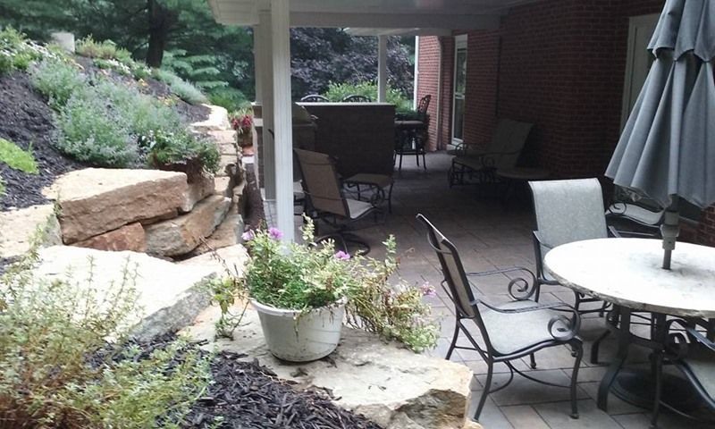 Stone patio with outdoor furniture, including chairs and table, under a covered area, next to a landscaped garden.