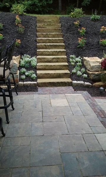 Stone steps lead up a hill, flanked by plants and dark mulch, from a patio.