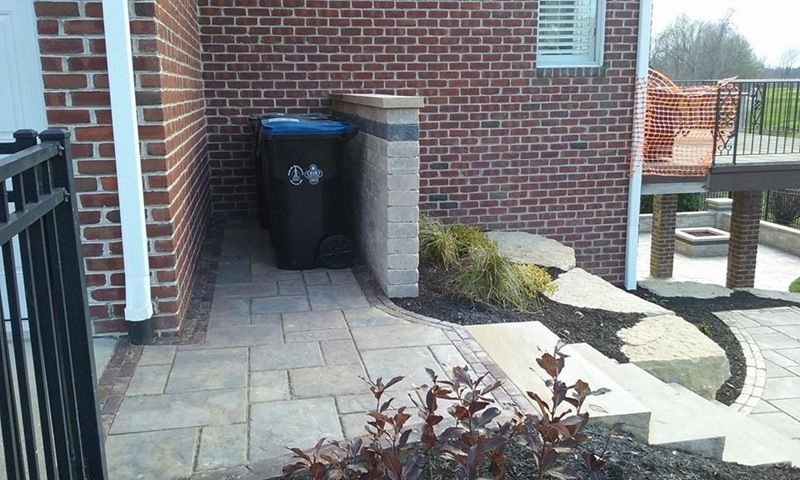 Trash can next to a brick wall and a paved walkway near a house and deck.