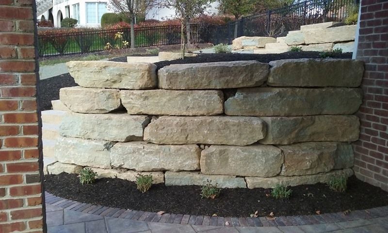 Stone retaining wall, curved, built with large, tan blocks, black mulch at base, brown brick wall on right.