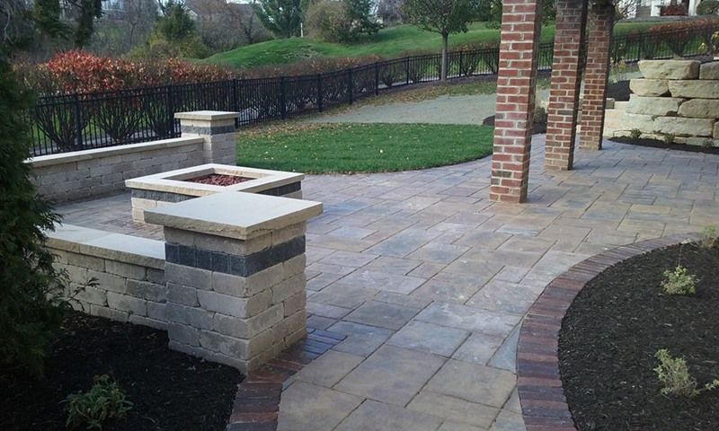 Patio with brick pillars, stone pavers, a fire pit, and lawn; black fence and shrubs in background.