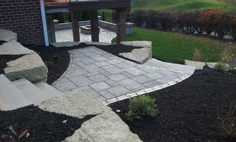 Stone patio with pathway and steps; surrounded by black mulch, greenery, and a grassy hill.