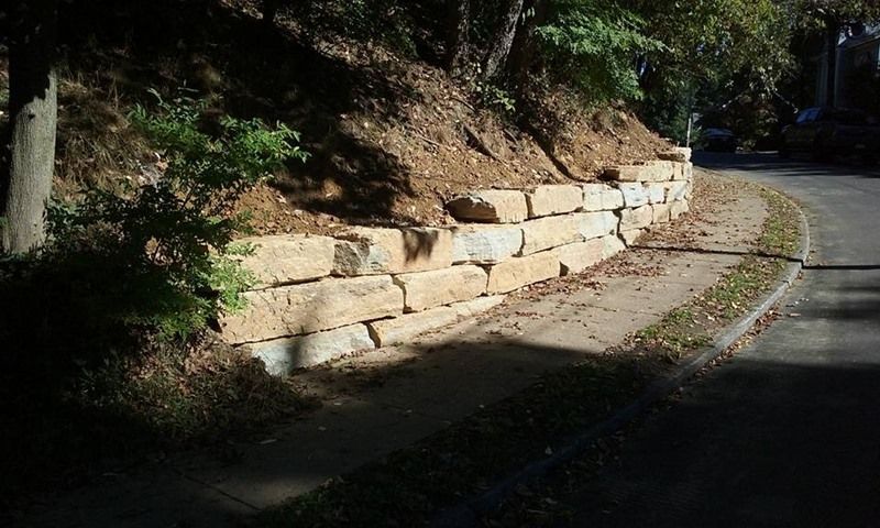 Stone retaining wall beside a sidewalk and road, with hillside above.