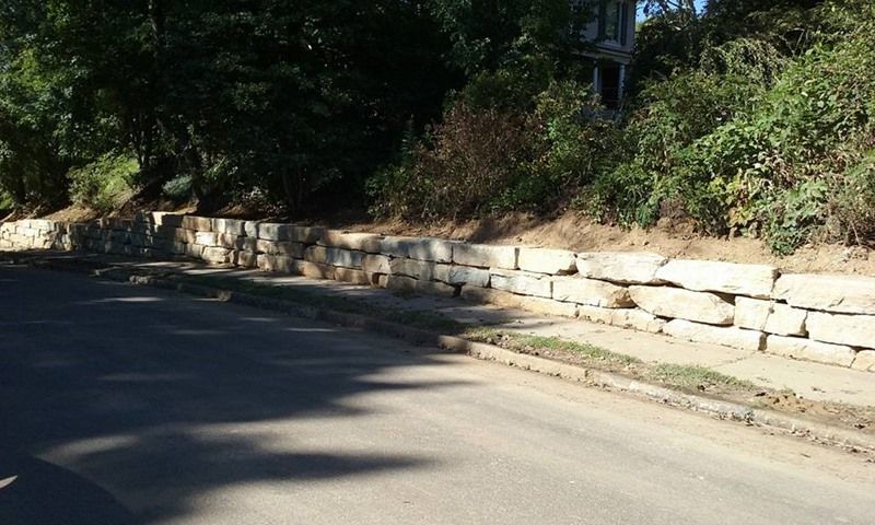 Stone retaining wall bordering a street with sidewalk, topped with dry grass; trees and a building in the background.