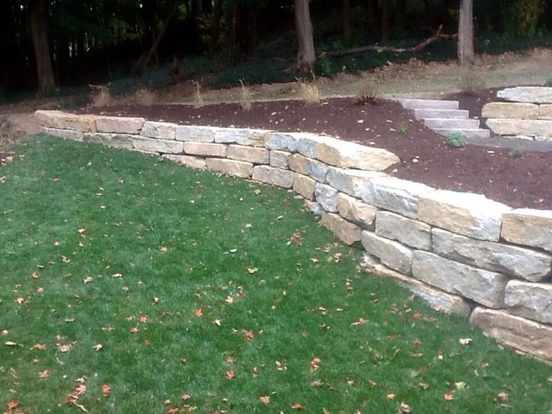 Stone retaining wall in a yard, with grass and soil. Steps lead upwards.
