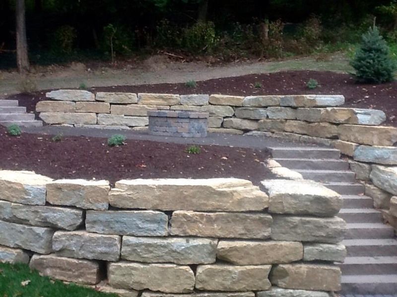 Stone retaining walls with steps leading to a fire pit area, surrounded by mulch and greenery.