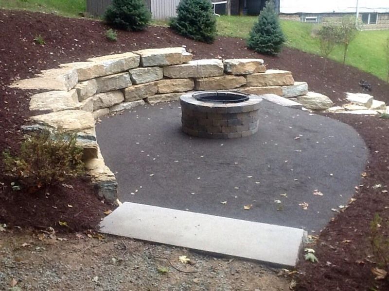Stone fire pit area with retaining wall, on a sloped yard with mulch and evergreen trees.