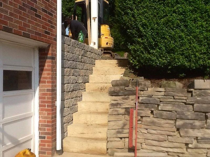 Stone staircase built into a retaining wall, leading to a small excavator at the top, next to a garage door.