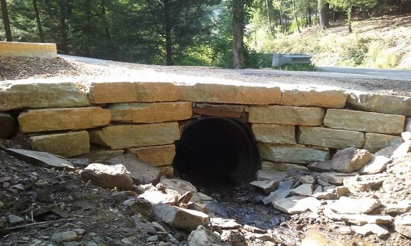 Stone culvert under a road with dark opening, water flowing out. Sunlight illuminates the scene.