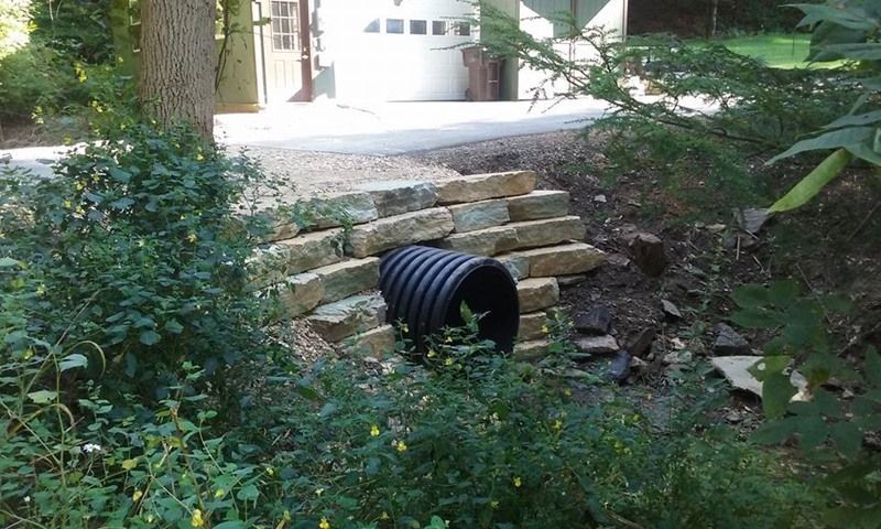 Black culvert pipe under a roadway supported by stone blocks, surrounded by greenery.