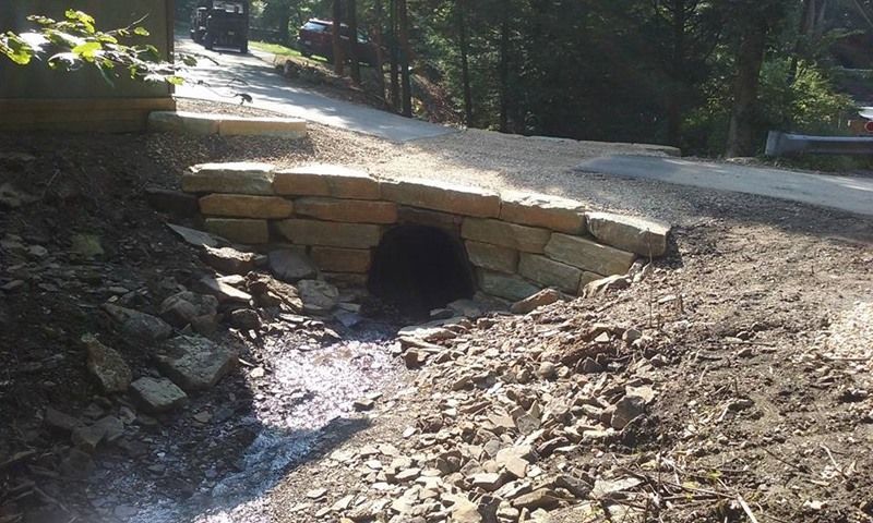 Stone bridge over a stream on a gravel road. Dark tunnel under the bridge, trees in the background.