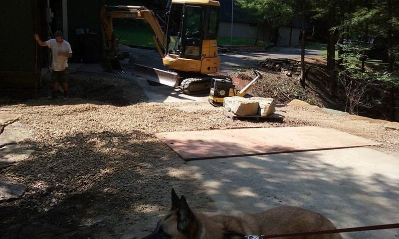 Man and dog watch excavator on a gravel and concrete surface next to a building; construction in progress.