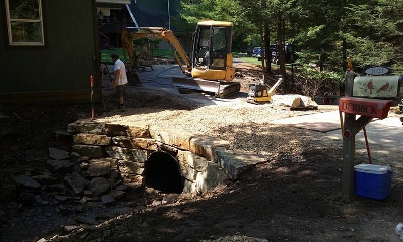Construction site: stone culvert under a gravel path. Excavator and worker visible near house and mailbox.
