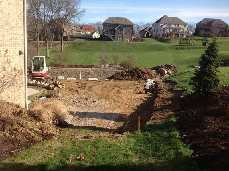 Construction site with excavator, dirt, and landscaping in progress; suburban houses in background.