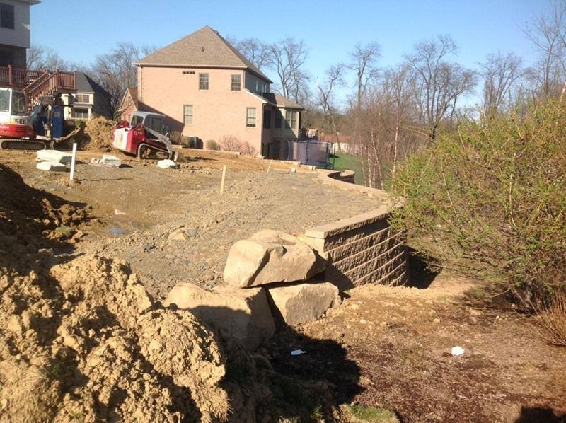 Construction site with retaining wall, dirt piles, small excavator, and house in the background.