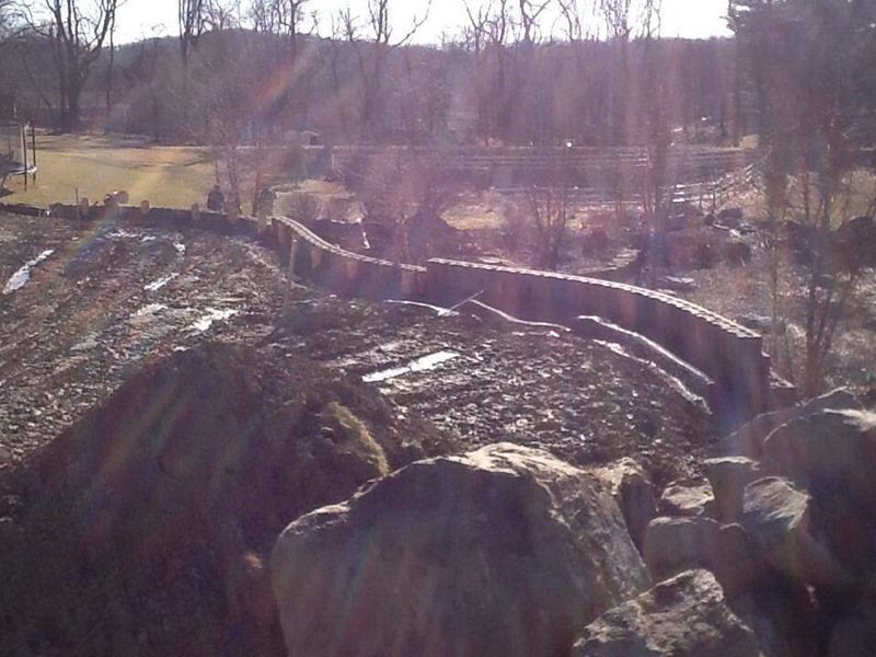 A dry, rocky landscape with a curved stone structure. Sunlight streams through bare trees.