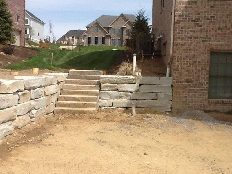 Stone retaining walls with stairs leading up to a grassy hill, next to a brick building.