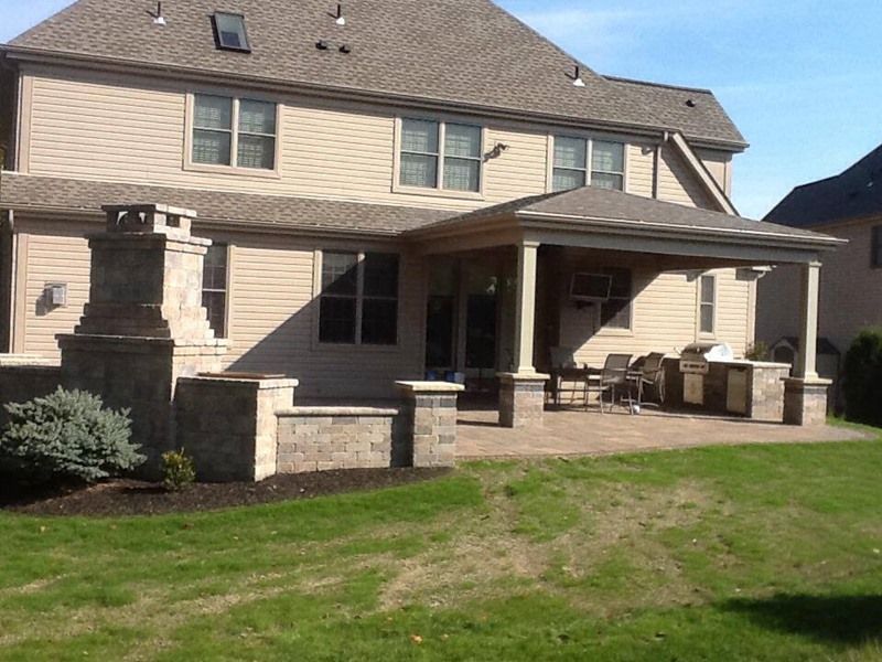 Backyard patio with fireplace and covered seating area, beige siding, stone accents, and green lawn.