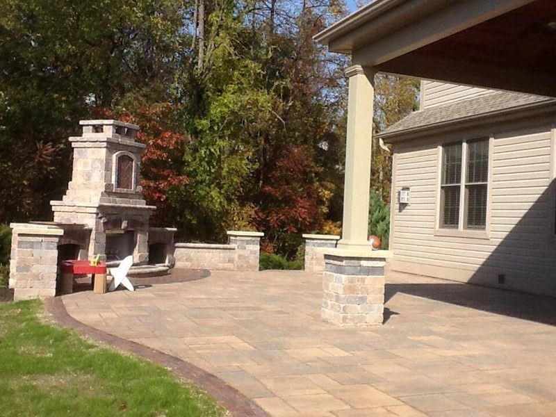Stone patio with fireplace and pillars next to a house with trees in the background.
