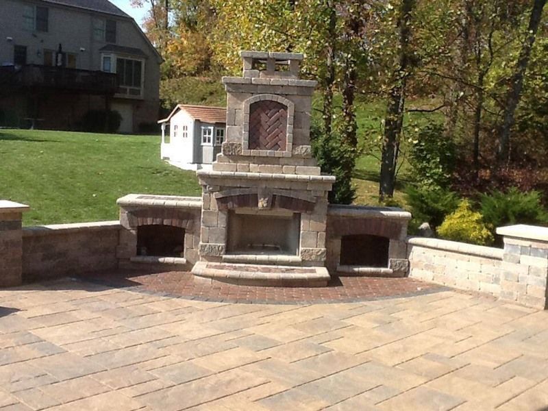 Outdoor stone fireplace on a brick patio with storage alcoves. A shed and grassy yard are in the background.