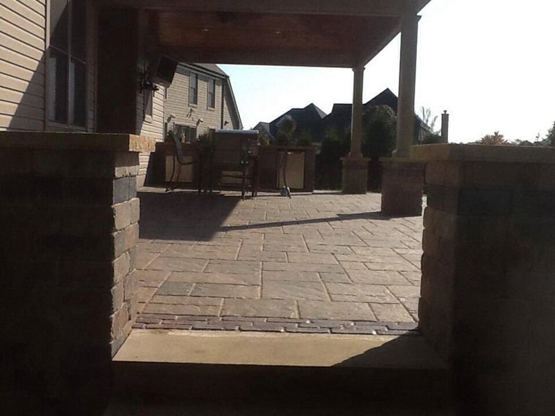 Stone patio with a covered seating area; view toward a house and trees.