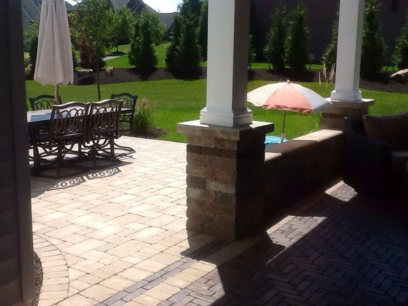 Patio with brick pavers, table and chairs, umbrella, pillars, and green lawn.