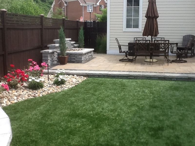 Patio with outdoor furniture, fire pit, and garden, featuring green lawn, beige paving, and a brown umbrella.