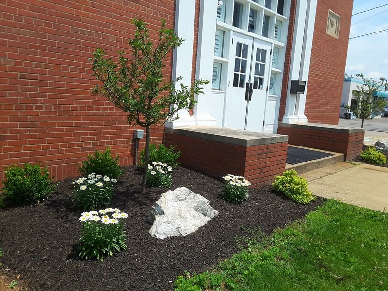 Building entrance with brick facade and landscaped flower bed; tree, flowers, and rock in mulch.