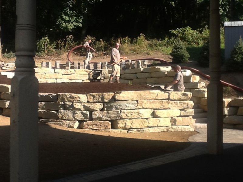 Three people working on stone wall construction outdoors. One person rolls a pipe.
