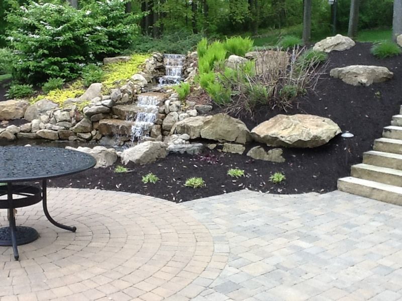 Backyard patio with waterfall feature, stone steps, table, and dark mulch.