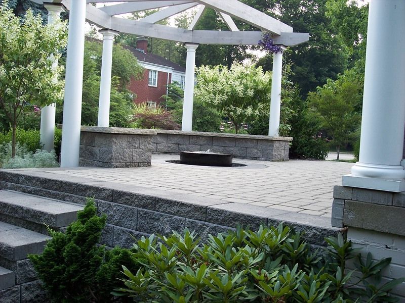 Stone patio with white pergola, seating area, and fountain. Trees and a red brick house are in the background.
