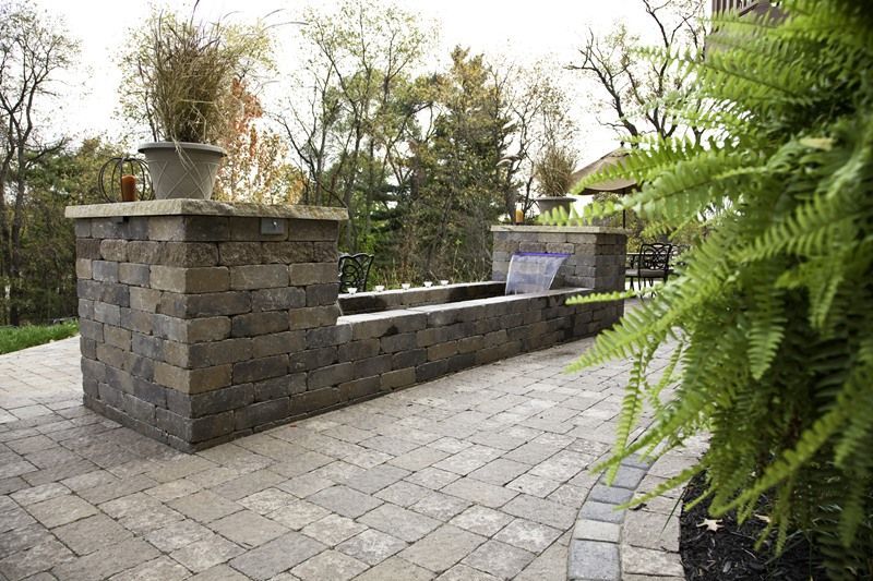 Stone wall with water feature, brick patio, and green plants in an outdoor setting.