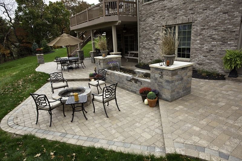 Stone patio with fire pit and seating, leading to tiered water feature and upper deck. Brick house in background.