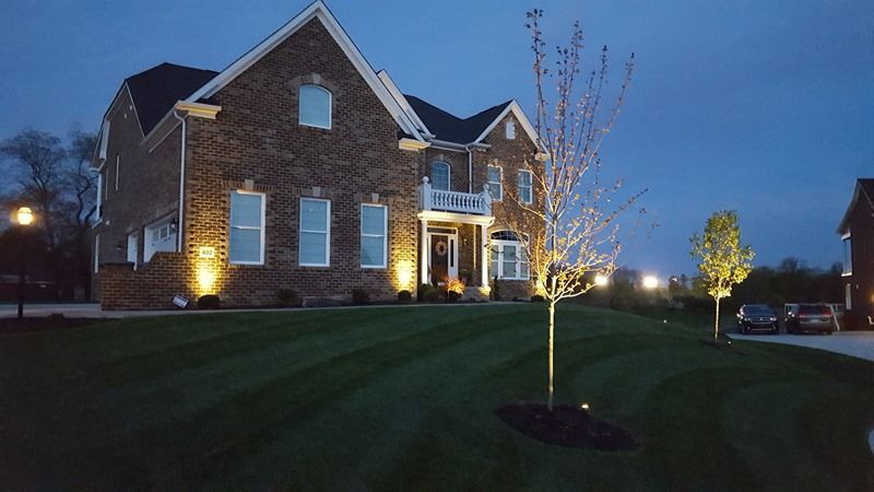 Two-story brick house illuminated by ground lights, set on a manicured lawn under a dusky sky.