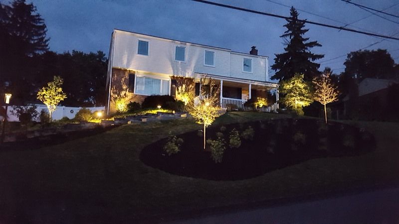 Two-story house illuminated with outdoor lighting on a dark night. Trees and landscaping in front.