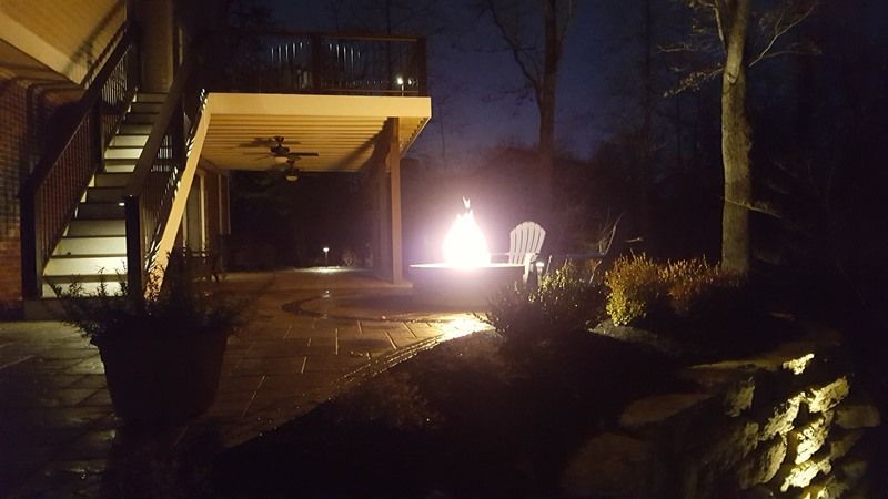 Nighttime outdoor scene: Deck with stairs, fire pit with two chairs, and landscape lighting.