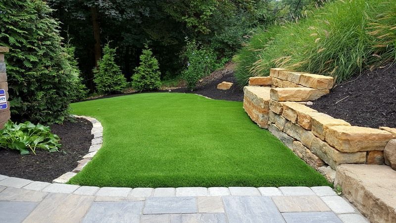 A patch of green artificial turf in a backyard, edged by stone and dark mulch, stone wall on right.