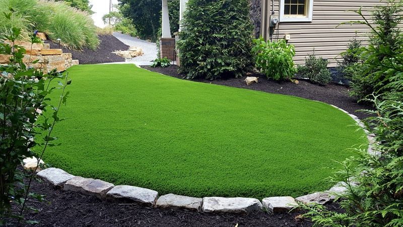 A small patch of bright green artificial turf surrounded by dark mulch and stone edging in a backyard.