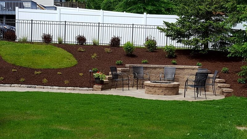 A backyard patio with a fire pit, seating, and a retaining wall with landscaping and a white fence.