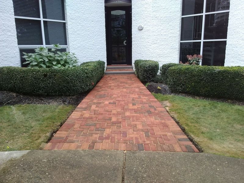 Brick pathway leading to a black door, flanked by green hedges and windows on a white stucco building.