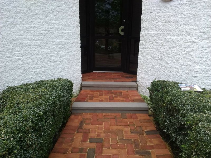 Brick walkway and steps leading to a dark door, flanked by green hedges, white building walls.