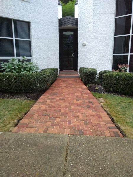 Red brick pathway leading to a black door, flanked by hedges, between white buildings.