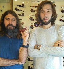 Two men with beards are standing next to each other holding pipes.