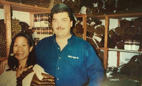 A man and a woman are posing for a picture in front of a shelf full of cigars
