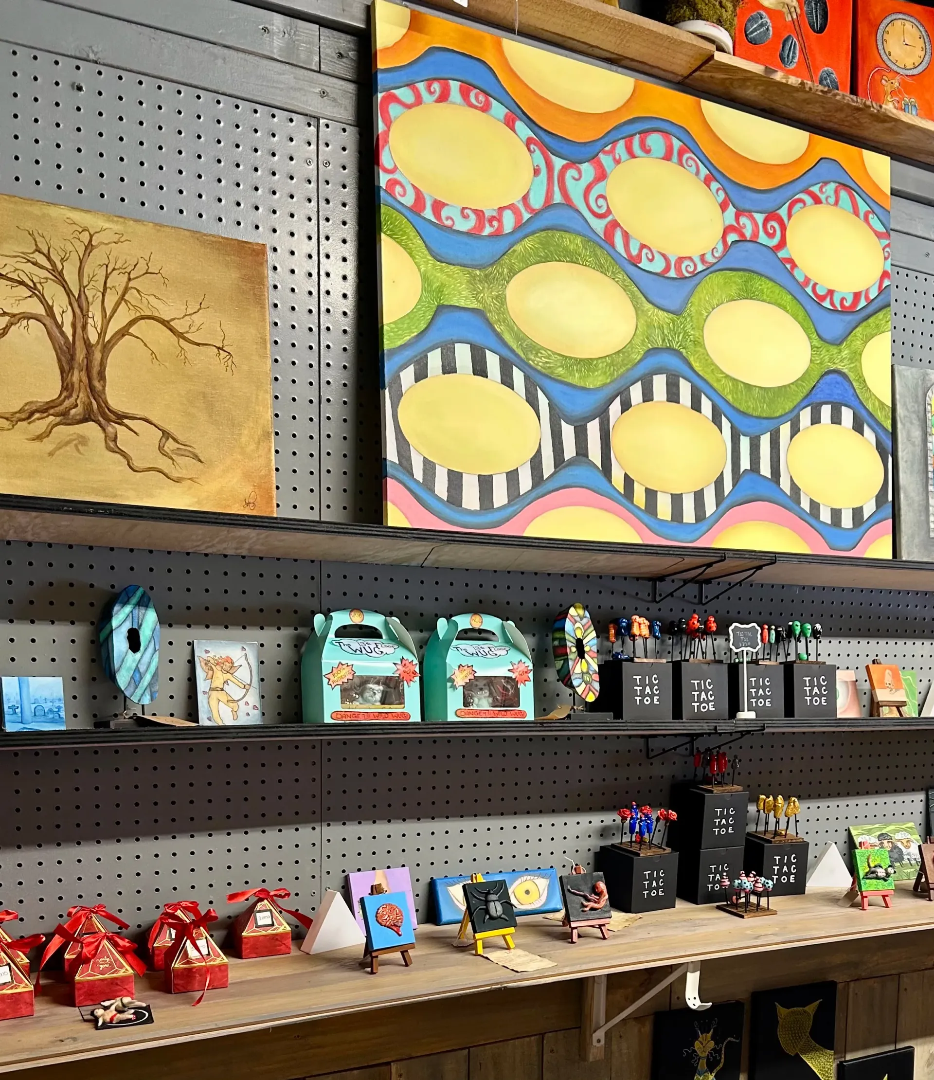 Shelves in a shop display paintings, colorful boxes, small framed art, and miniature models against a gray pegboard wall.