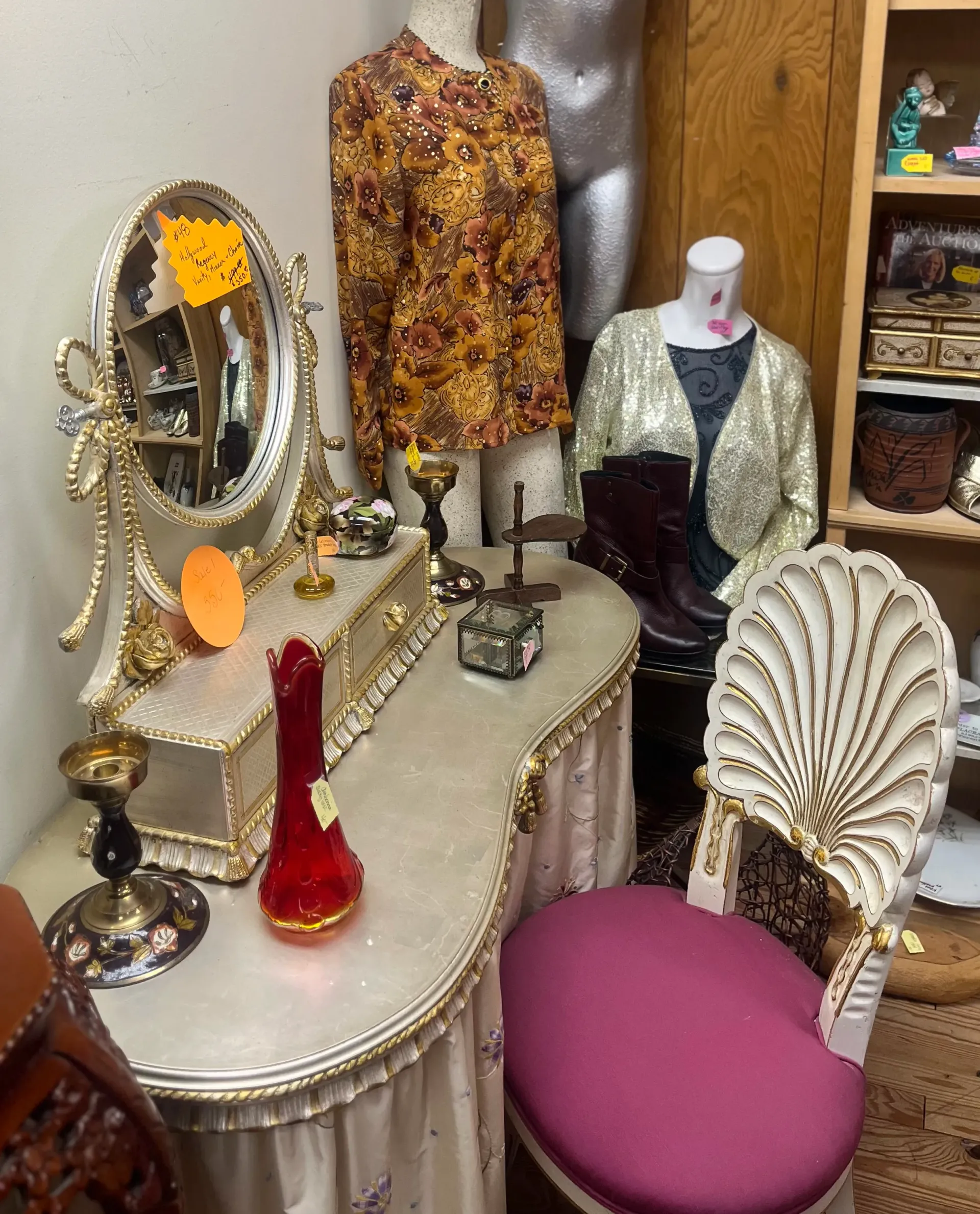 A vanity table with a mirror, red vase, and seashell-back chair in a shop, flanked by mannequins wearing vintage clothing.