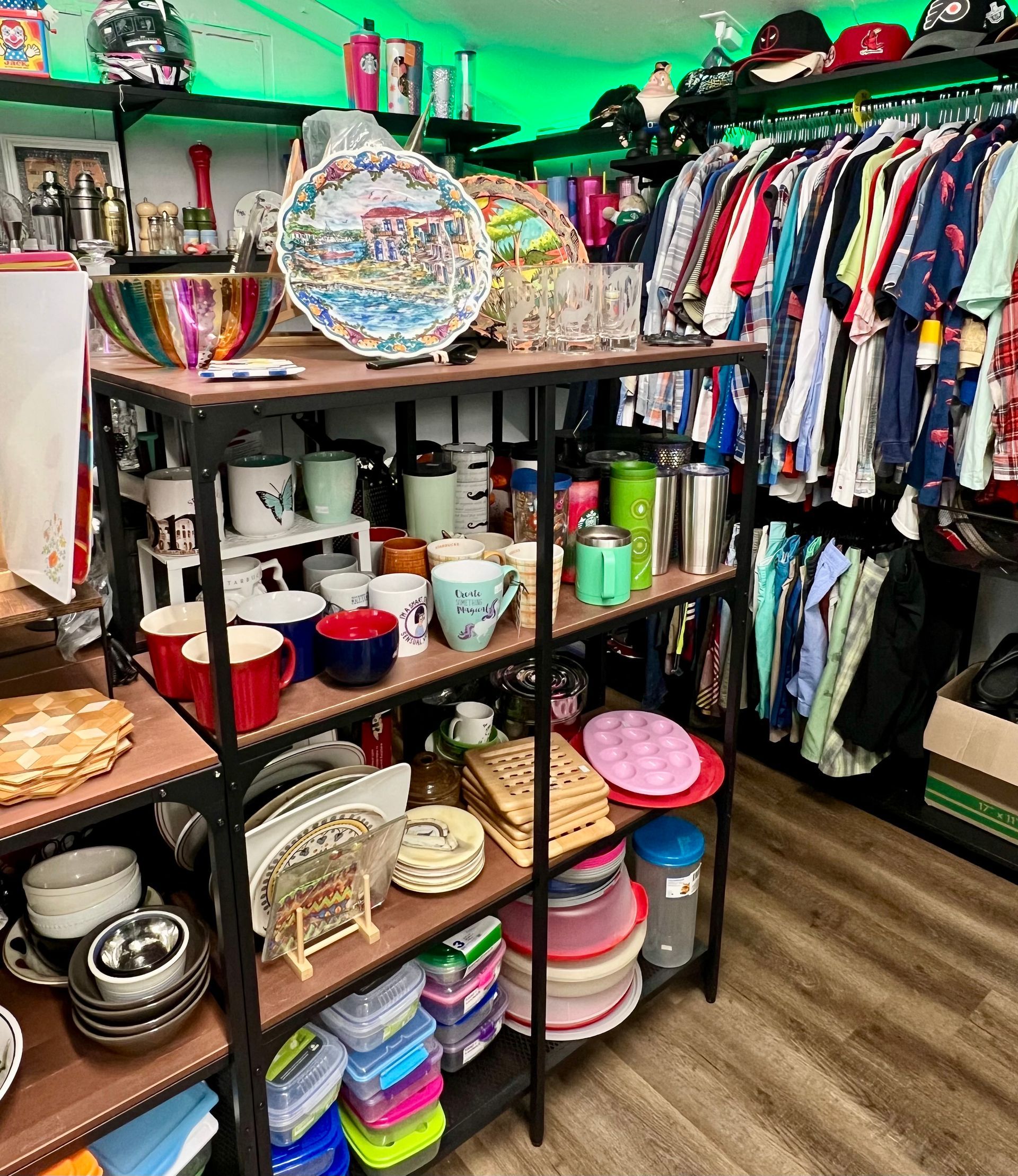 A metal shelving unit filled with various mugs, bowls, and storage containers, standing next to a rack of hanging shirts.