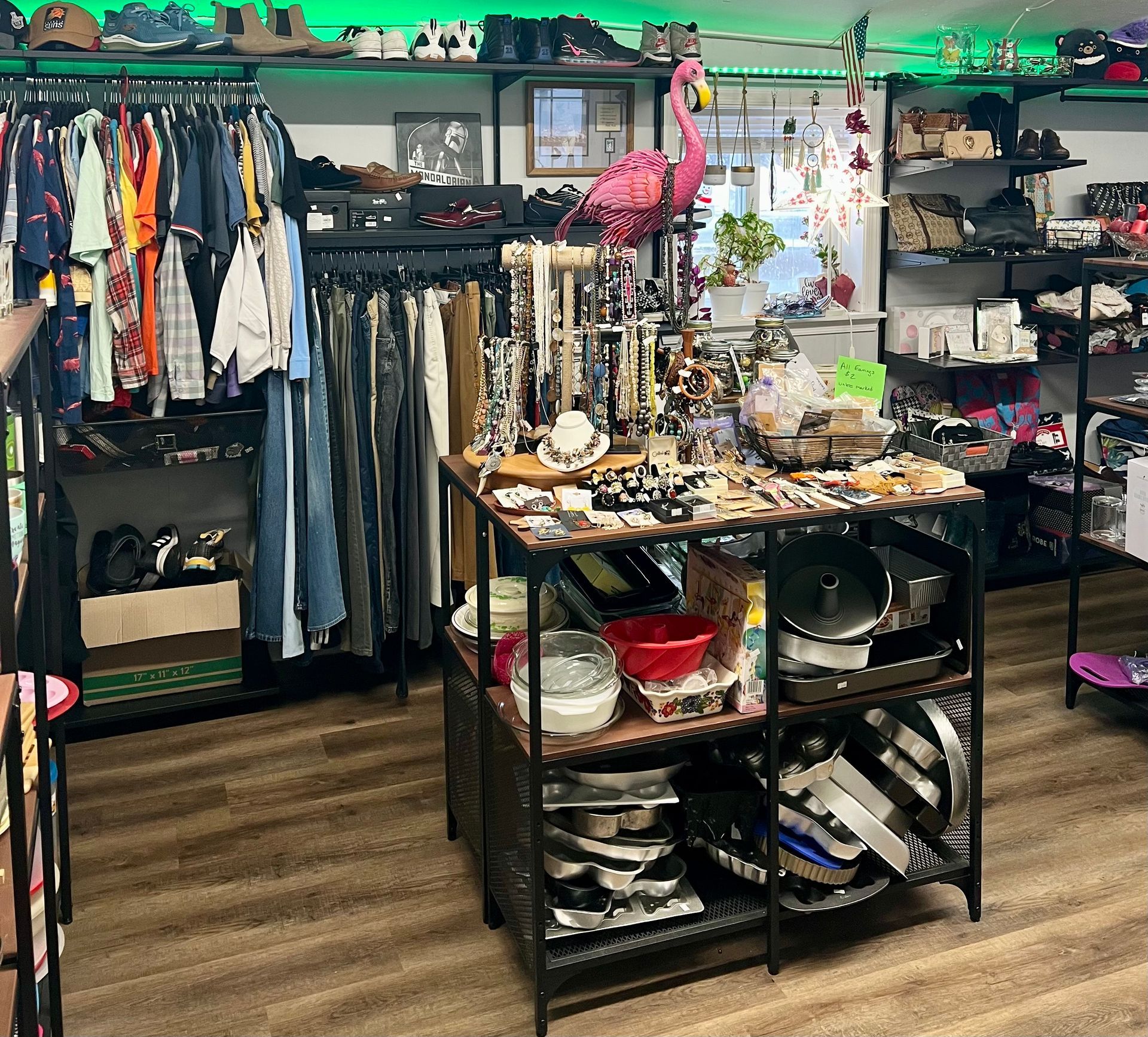 A cluttered thrift store interior with clothing racks, a central display shelf filled with housewares, and a pink flamingo.