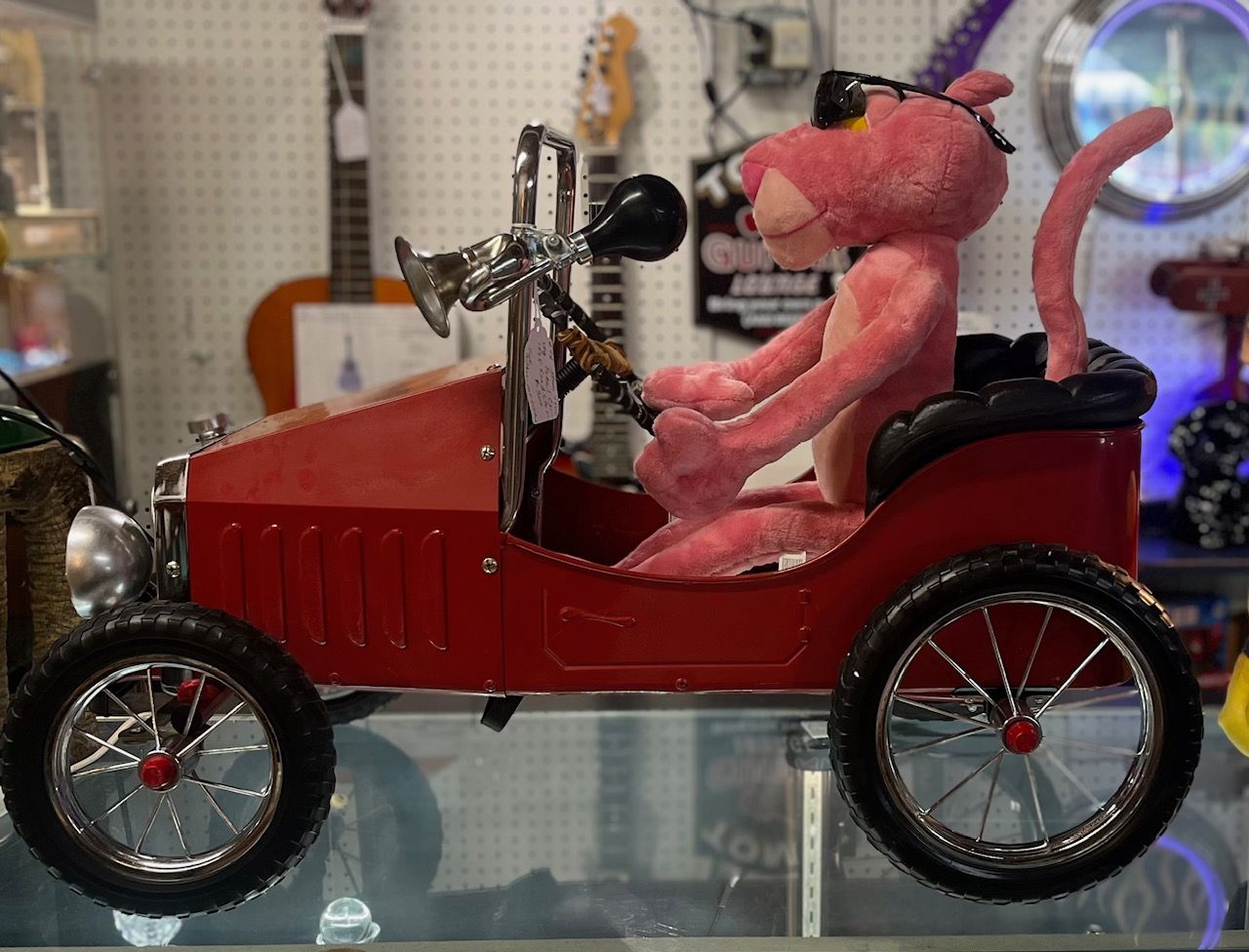 Pink Panther toy in a red vintage car, wearing sunglasses, indoors.