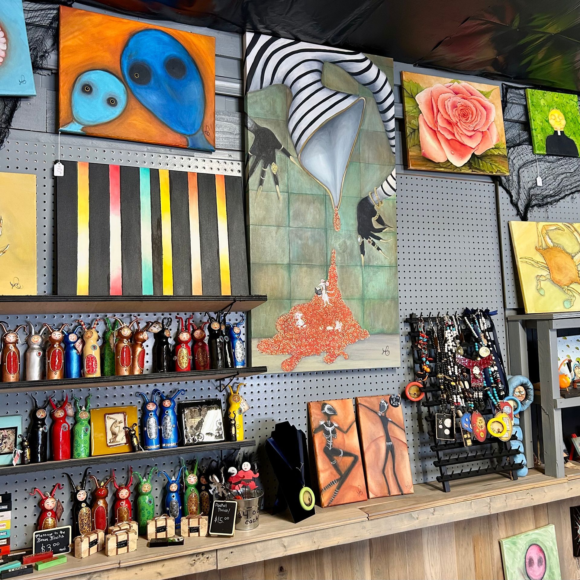 A shop wall displaying various paintings and rows of small, colorful handmade figurines on wooden shelves.
